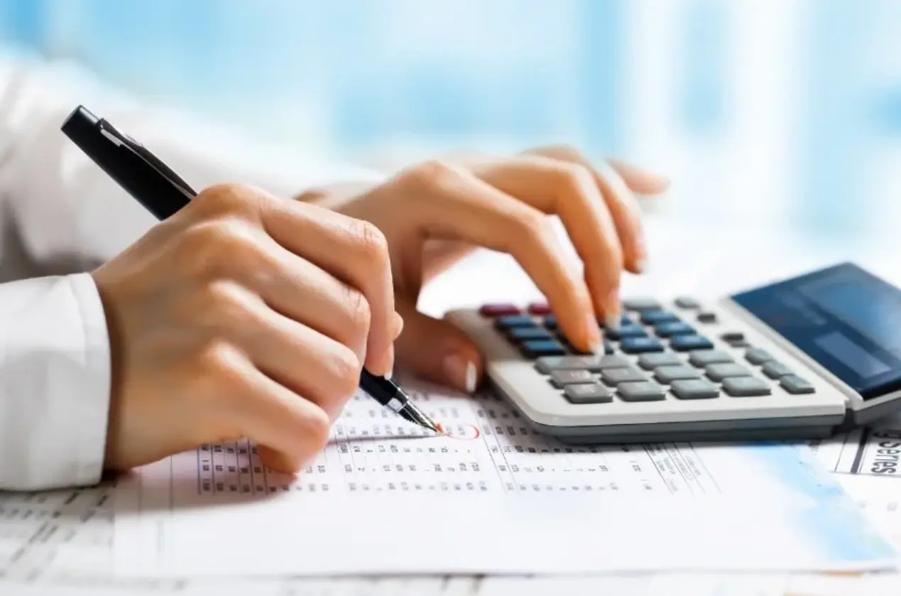 A person engages with a calculator and pen, recording information on paper while consulting a debit credit cheat sheet