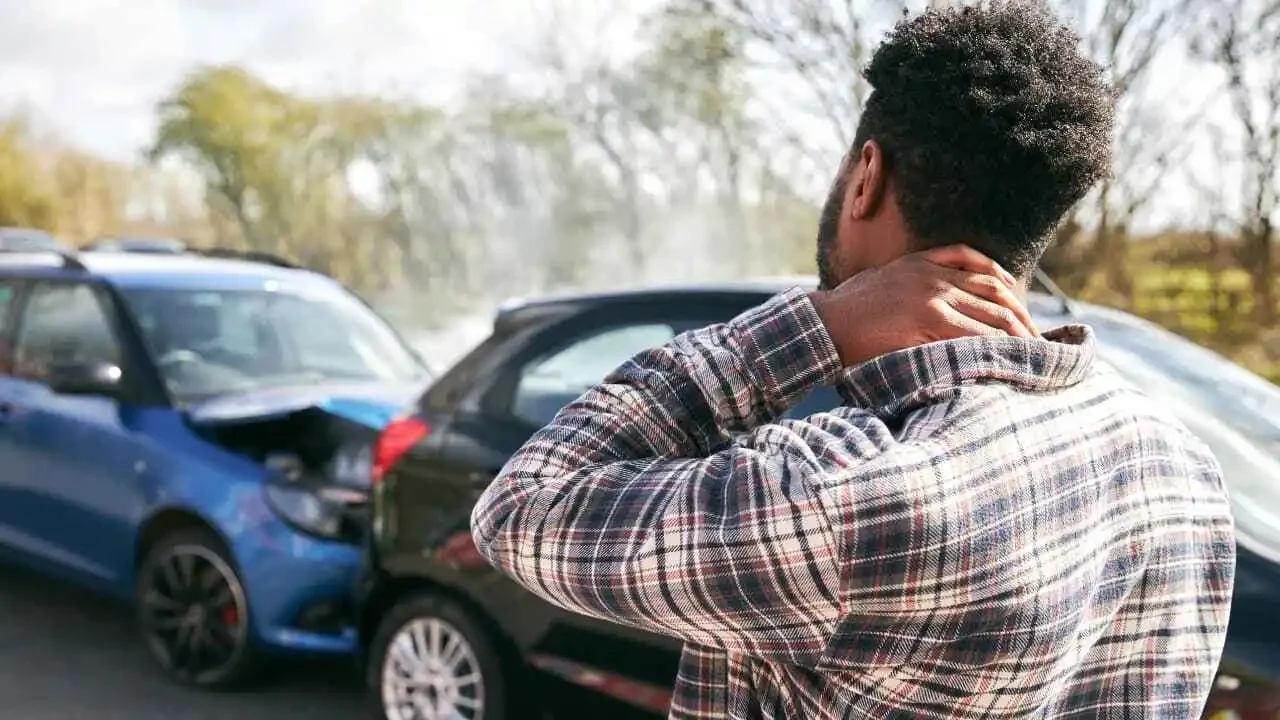 A man holds his neck in pain while staring at his car, symbolizing struggles after a car accident without insurance.