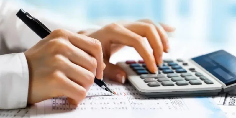 A person engages with a calculator and pen, recording information on paper while consulting a debit credit cheat sheet